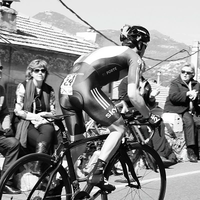 a black and white image of a male biker biking along the road next to a crowd of onlookers